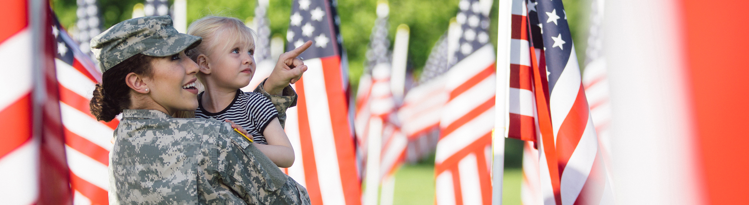 A woman in a military uniform is smiling, carrying a little girl, and pointing to something in the distance amidst a sea of American flags. 